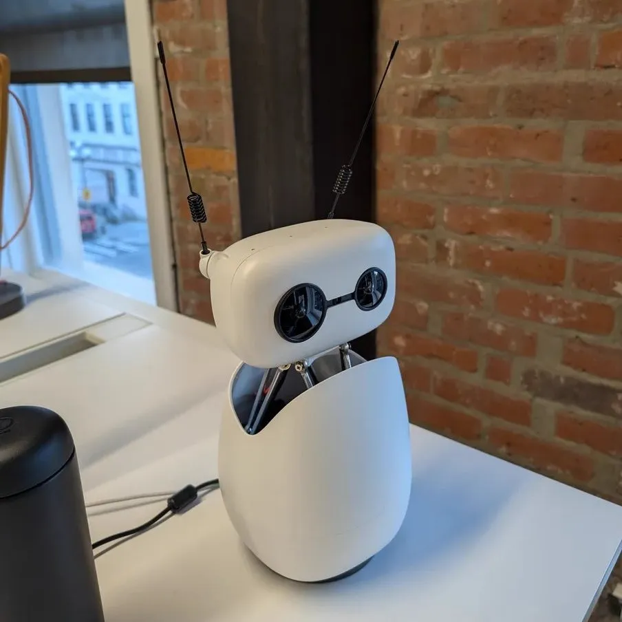 Reachy Mini, a small white robot with two round black eyes and two thin antennas, sitting on a white desk in front of a brick wall.